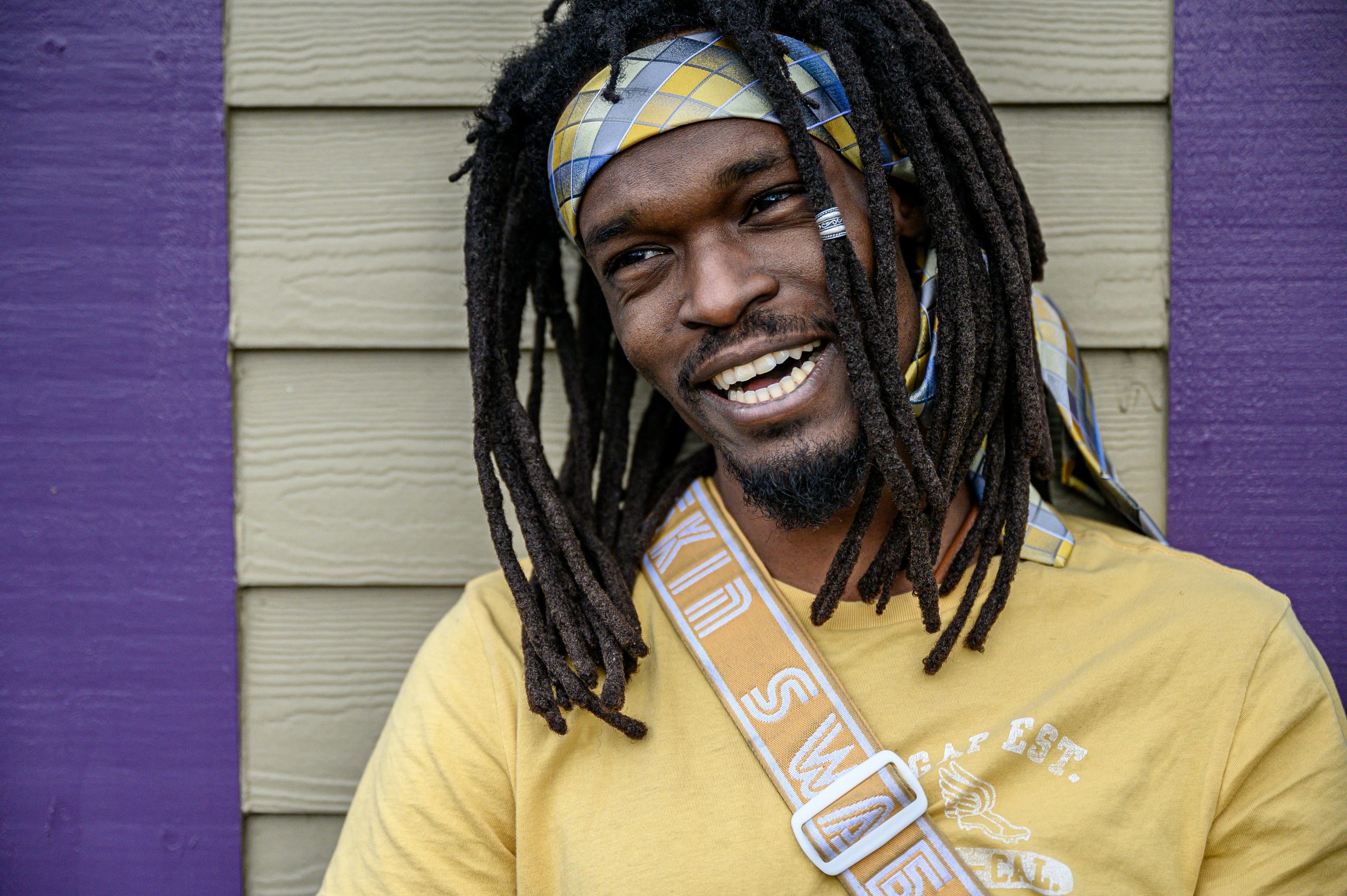 Portrait of a smiling man with dreadlocks against a colorful background wearing a yellow shirt.